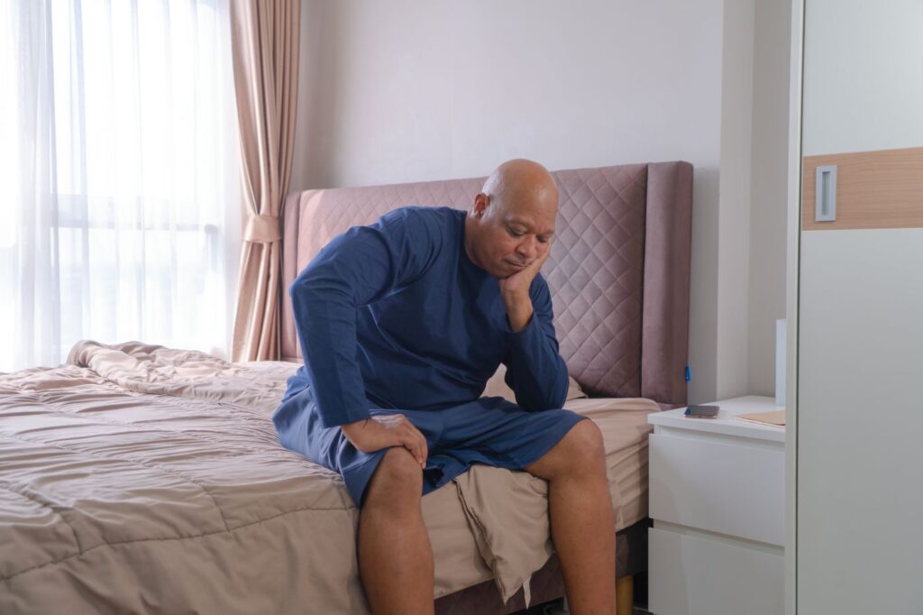 Homem com expressão preocupada, sentado em sua cama e com a mão no rosto, pensa se próstata aumentada é grave