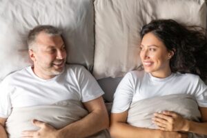 Casal sorrindo deitado na cama e trocando olhares, ilustrando a vida sexual após cirurgia de próstata e a retomada da intimidade no relacionamento.