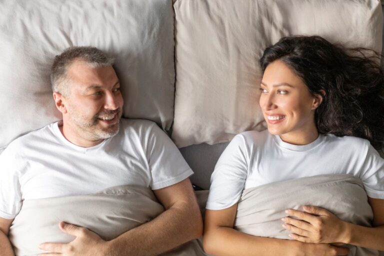 Casal sorrindo deitado na cama e trocando olhares, ilustrando a vida sexual após cirurgia de próstata e a retomada da intimidade no relacionamento.